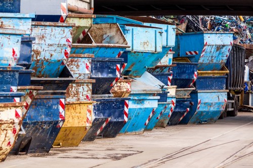 A row of large industrial metal skips are lined up along a concrete surface, each with a rectangular shape and built from steel, showing signs of rust and wear with chipped paint in shades of blue, yellow, and black. The skips are positioned side by side, some slightly tilted or overlapping, and feature red and white striped reflective safety stickers on their edges. In the background, stacks of additional waste materials and possibly a truck or storage area are partially visible, indicating an outdoor waste management or rubbish collection site. The scene is illuminated by natural daylight, highlighting the textures of the rusted and weathered surfaces, consistent with commercial waste removal activities in an urban environment, such as Finsbury Park area. Commercial Waste Removal Finsbury Park operates in the surrounding postcode area, providing waste disposal services for businesses dealing with industrial or large-scale rubbish.