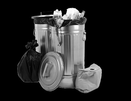 In the foreground of the image, there is a large, cylindrical metallic rubbish bin with a closed lid, which is partially covered by a black plastic bag filled with waste, tied at the top. Next to the bin, on the right, lies a white plastic bag that appears crumpled and contains additional rubbish. To the left of the bin, a smaller black plastic bag is positioned upright, leaning against the side of the bin. The bin's surface has a reflective metal finish with visible vertical ridges along its body, and the lid is flat with a small handle in the centre. Inside the open top of the bin, various waste items are visible, including crumpled paper, plastic bottles, and possibly some packaging materials. The background is solid black, providing a stark contrast that emphasizes the objects and gives the scene a clean, professional appearance suitable for a rubbish removal service website in Finsbury Park, London, highlighting the company's fulfilment of local waste disposal needs.