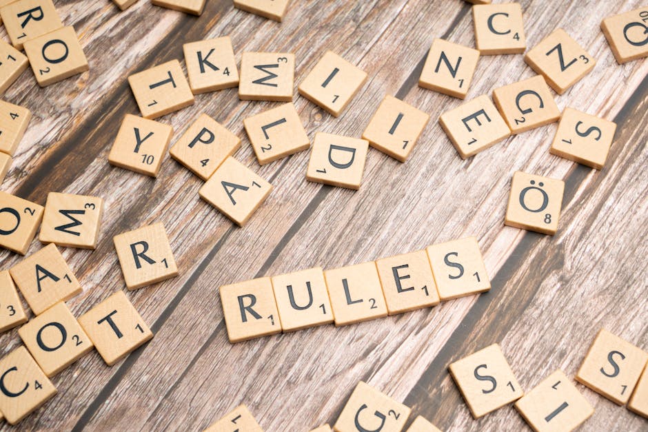 A close-up photograph features numerous light wooden Scrabble tiles with black lettering and small numbered score indicators, scattered randomly across a weathered wooden surface with visible grain an