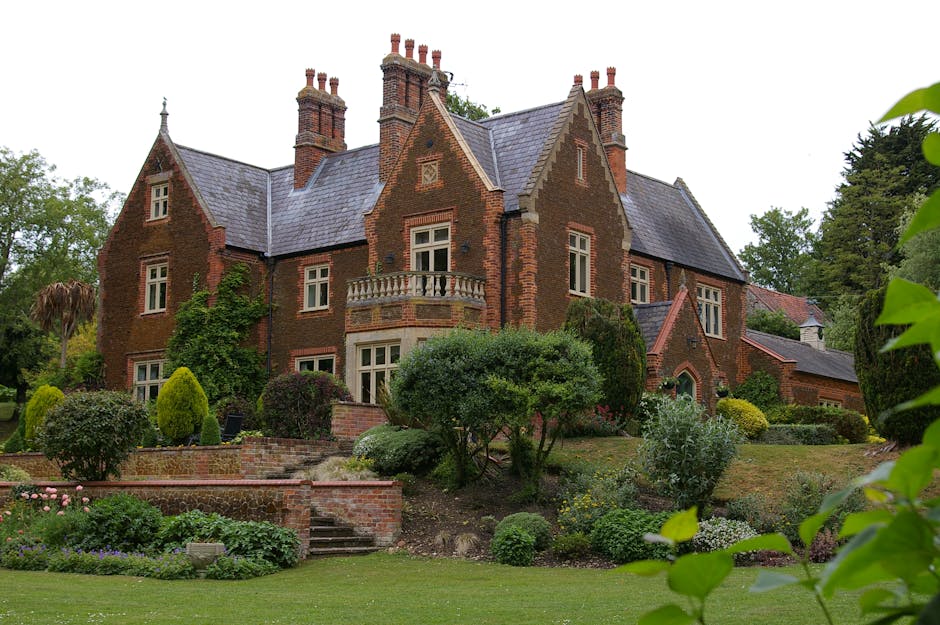 A historic multi-story building constructed from warm-toned sandstone features Gothic-style windows with intricate leaded glass panes and pointed arches, set amidst a landscaped garden with lush green