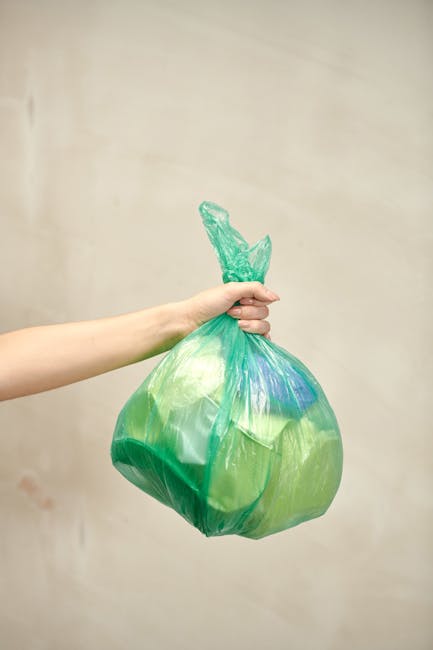 A person wearing a white t-shirt and transparent gloves is holding a white plastic bin labeled 'PLASTIC' in black letters. The bin contains a black garbage bag partially visible with an empty, clear p